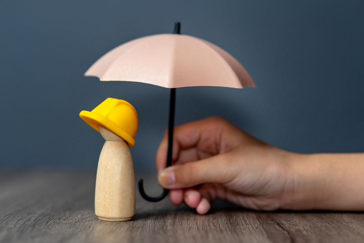 Nahaufnahme einer Hand, die einen Regenschirm über eine Holzpuppe mit gelbem Schutzhelm hält.