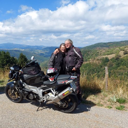 Annabell und ihr Mann stehen vor Motorrad, er legt den Arm liebevoll um ihre Schulter, Hügel und blauer Himmel im Hintergrund.
