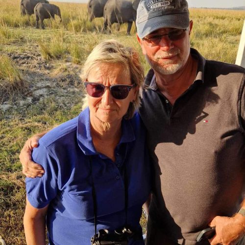 Christine und ihr Mann blicken in die Kamera, stehen an einem Geländer, im Hintergrund ist eine Elefantenherde zu sehen.
