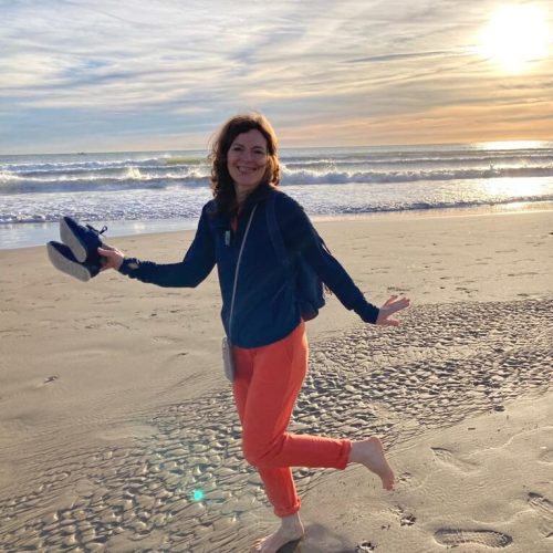 Lilia steht barfuß auf einem Bein am Strand im Sonnenuntergang, hält ihre Schuhe in einer Hand.