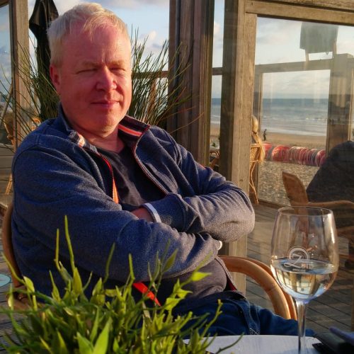 Peter sitzt mit verschränkten Armen und geschlossenen Augen im Außenbereich eines Restaurants, im Hintergrund Sand und Meer.