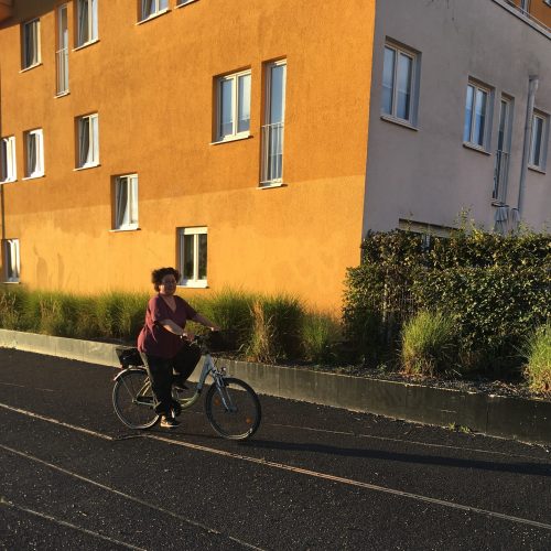 Sabine fährt Fahrrad in der Abenddämmerung vor einem orangen Haus.