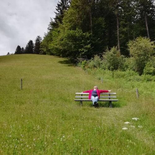 Antje sitzt auf einer Bank inmitten einer grünen Wiese am Waldrand.