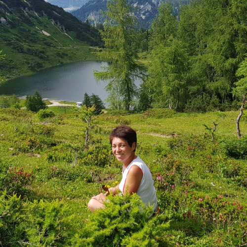 Hermine sitzt am Hang und schaut in die Kamera, im Hintergrund ein kleiner Bergsee, Berge und Tannenbäume.