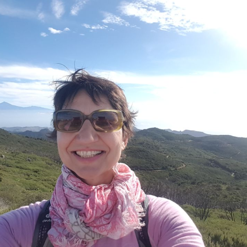 Barbara macht ein Selfie, sie lacht, im Hintergrund grüne Berglandschaft und blauer Himmel.