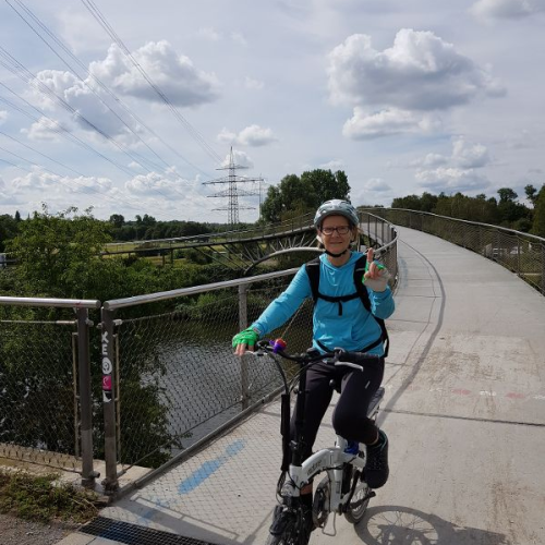 Erika radelt mit Helm und blauer Regenjacke auf die Kamera zu.