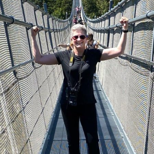 Gabi steht auf einer Hängebrücke, streckt die Arme nach oben und zeigt ihre Muskeln.
