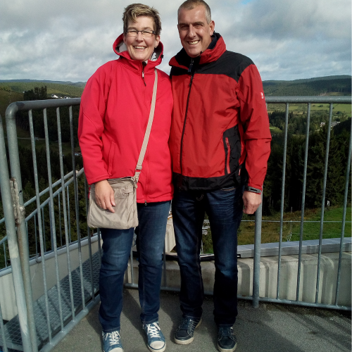 Sandra und ihr Mann stehen auf einer Aussichtsplattform, tragen rote Regenjacken, im Hintergrund grüne Hügel und bewölkter Himmel.